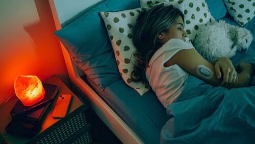 Young woman sleeping in bed next to her dog wearing a CGM on her upper right arm