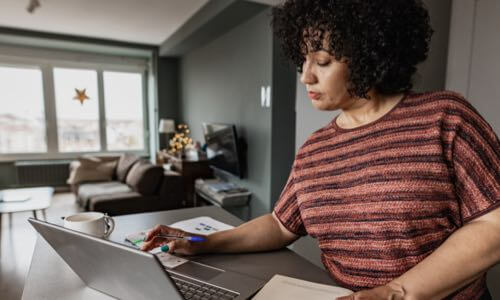Diverse female at a tall living room table viewing her open laptop with pen in hand & paper on table