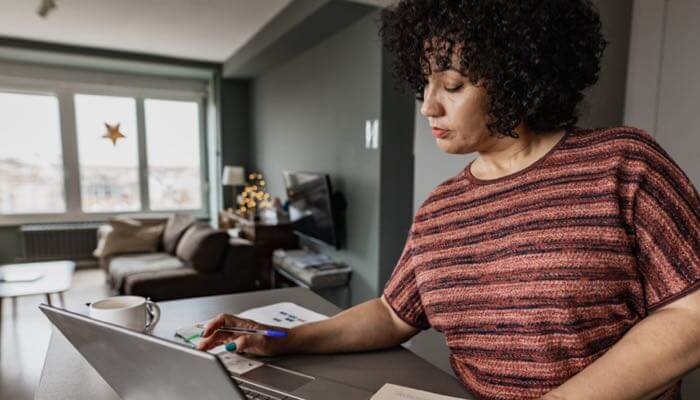 Diverse female at a tall living room table viewing her open laptop with pen in hand & paper on table