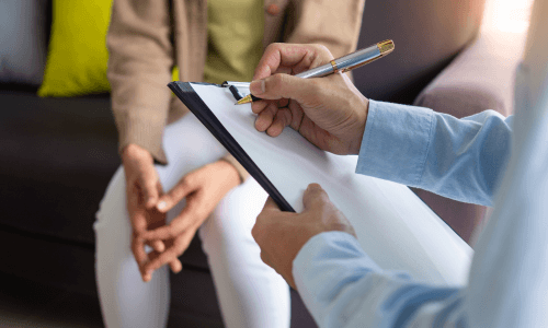 Close-up of a professional writing on a clipboard with a patient sitting in the background